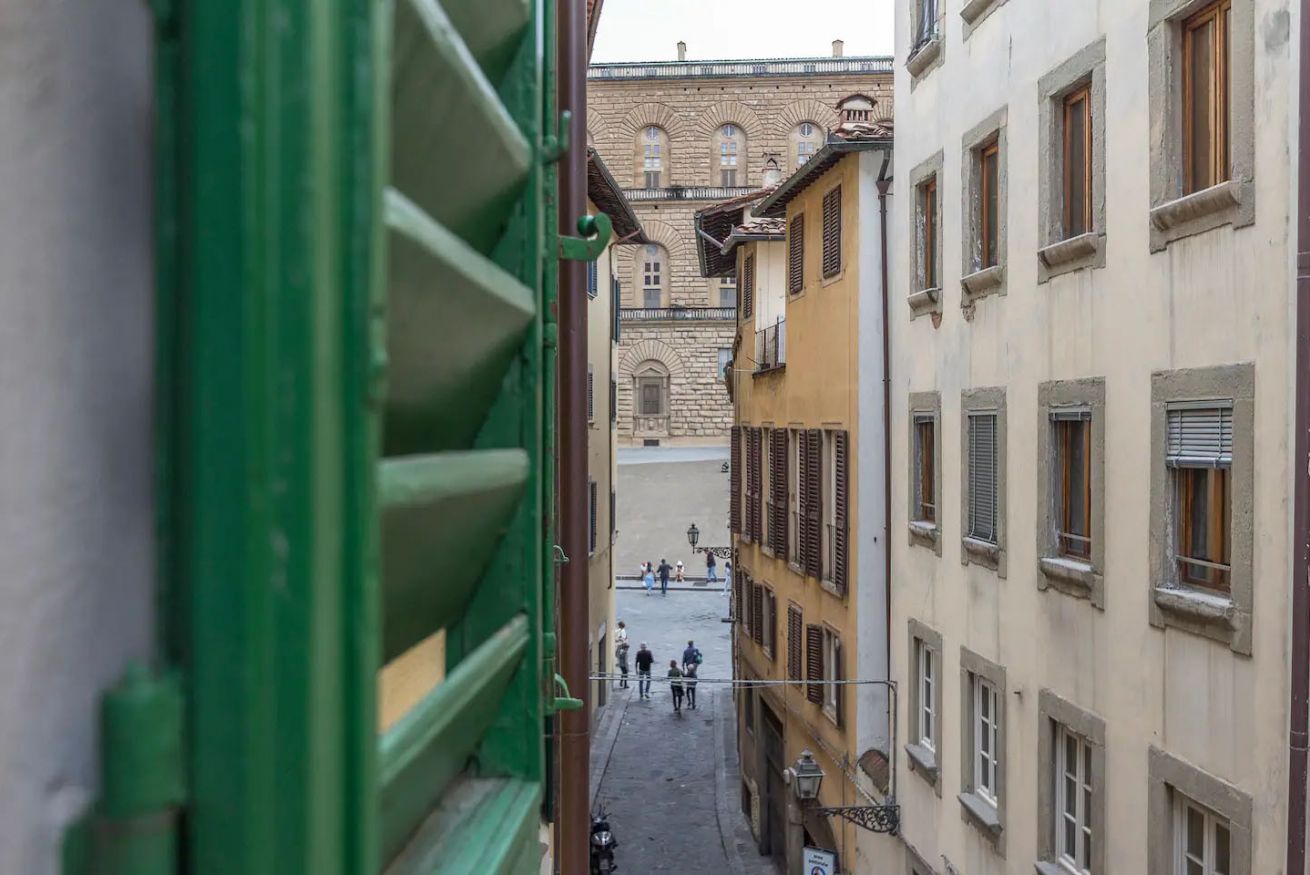 A narrow street with buildings on either side, a large stone building in the background, and a few people walking.