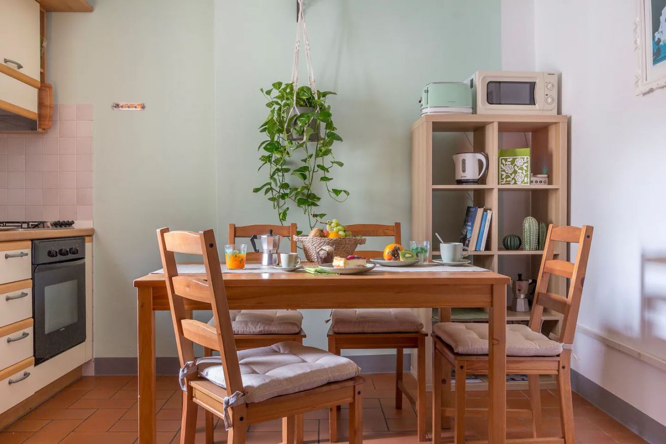 Une cuisine avec une table à manger, des chaises, une plante suspendue, un four à micro-ondes et une bibliothèque contenant des livres et des objets décoratifs.