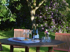 Een houten tafel buiten, compleet met fles, glazen, een bord eten en een kleine vaas bloemen.