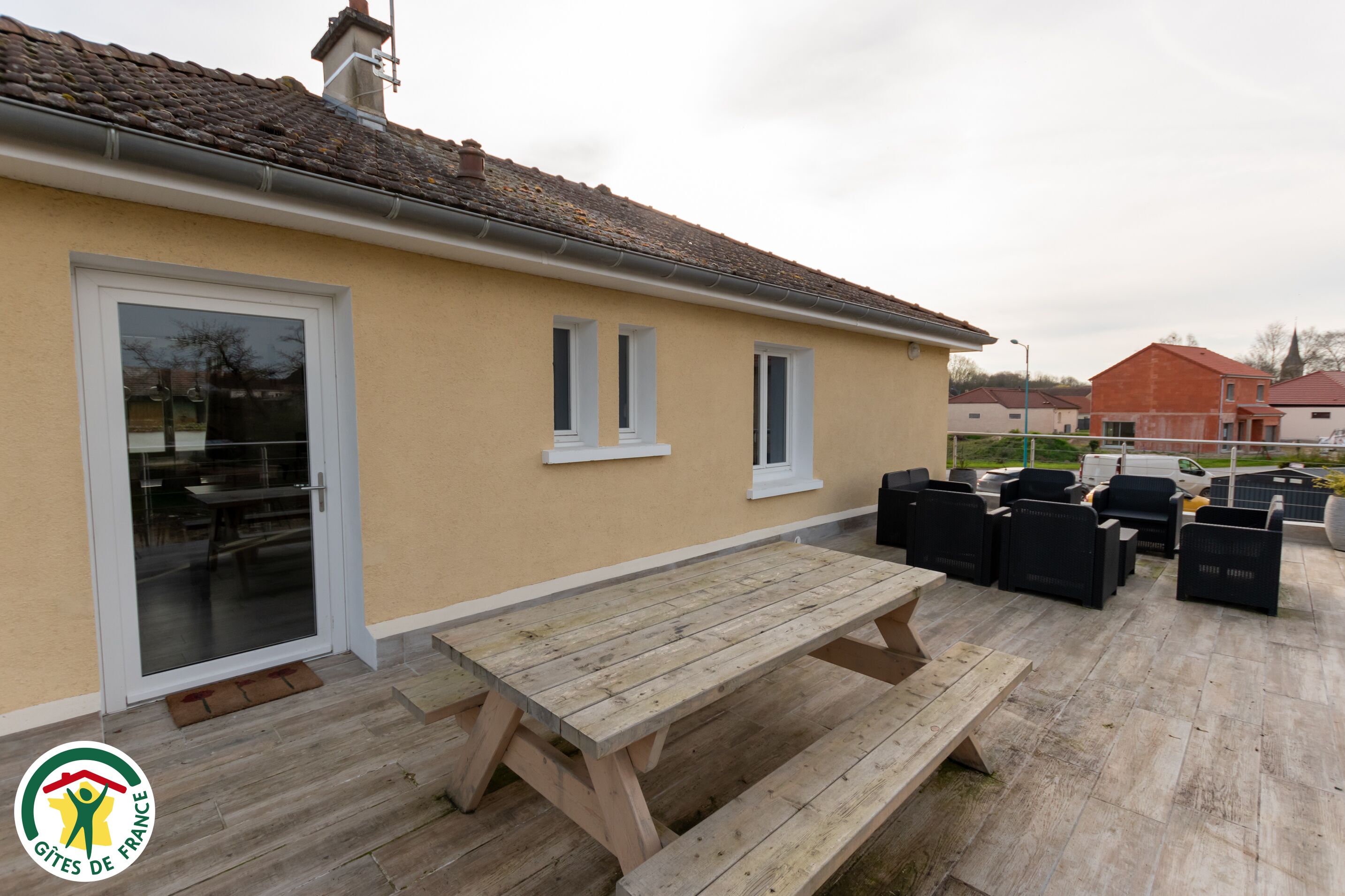 Un bâtiment à l’extérieur beige, doté d’un toit en tuiles et d’une terrasse en bois avec des sièges pour l’extérieur.