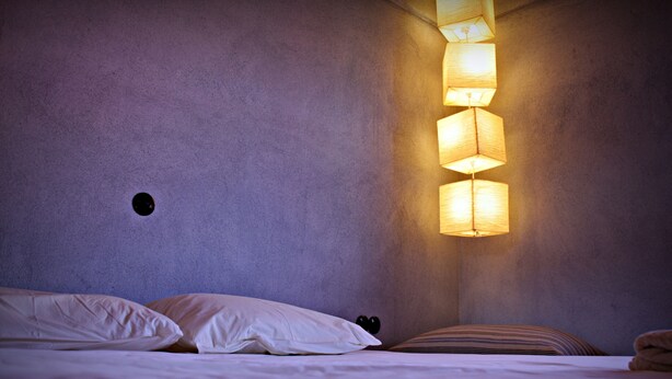 A bedroom with a bed, pillows, a wall-mounted light fixture, and a textured wall. - eumelia organic agrotourism farm (Evrotas)
