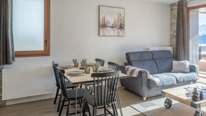 A modern living room with a dining area, featuring a grey sofa, wooden dining table, and a painting of a forest scene. - Bright apartment with terrace and parking in Alpe d'Huez (Huez)