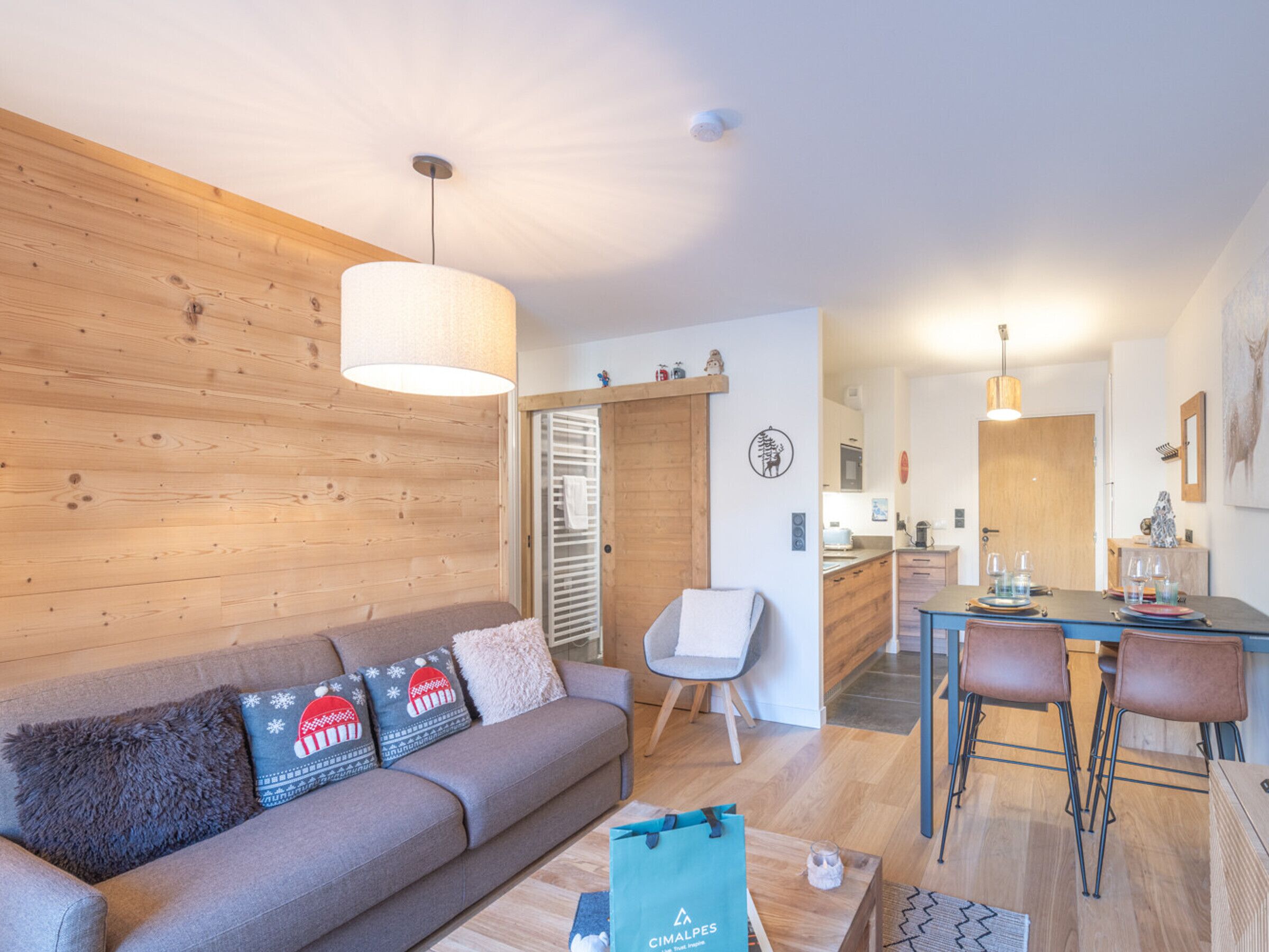 A modern living room with a wooden wall, a grey sofa with festive pillows, a coffee table, and a dining area with a wooden table and chairs.