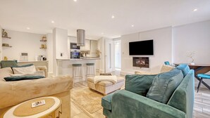 A modern living room with a sofa, ottoman, and a small round table. There is a kitchen area with a bar and a TV mounted on the wall. - The Luxe Hideaway - Stunning 2 bedroom apartment in Wanstead, East London (London)