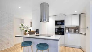 A modern kitchen with white cabinetry, a stainless steel range hood, and two blue bar stools. - The Luxe Hideaway - Stunning 2 bedroom apartment in Wanstead, East London (London)