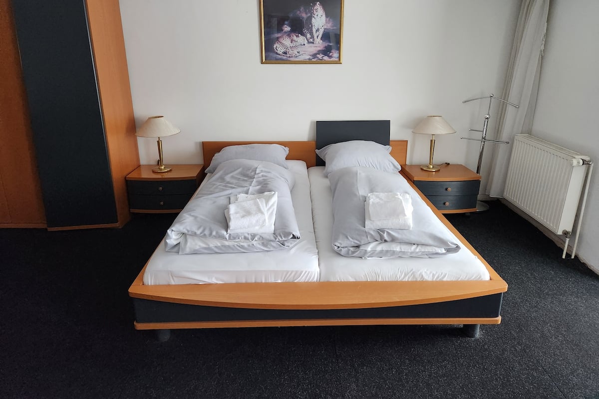 A double bed with white linens and towels, two bedside tables with lamps, a framed picture on the wall, and a radiator.