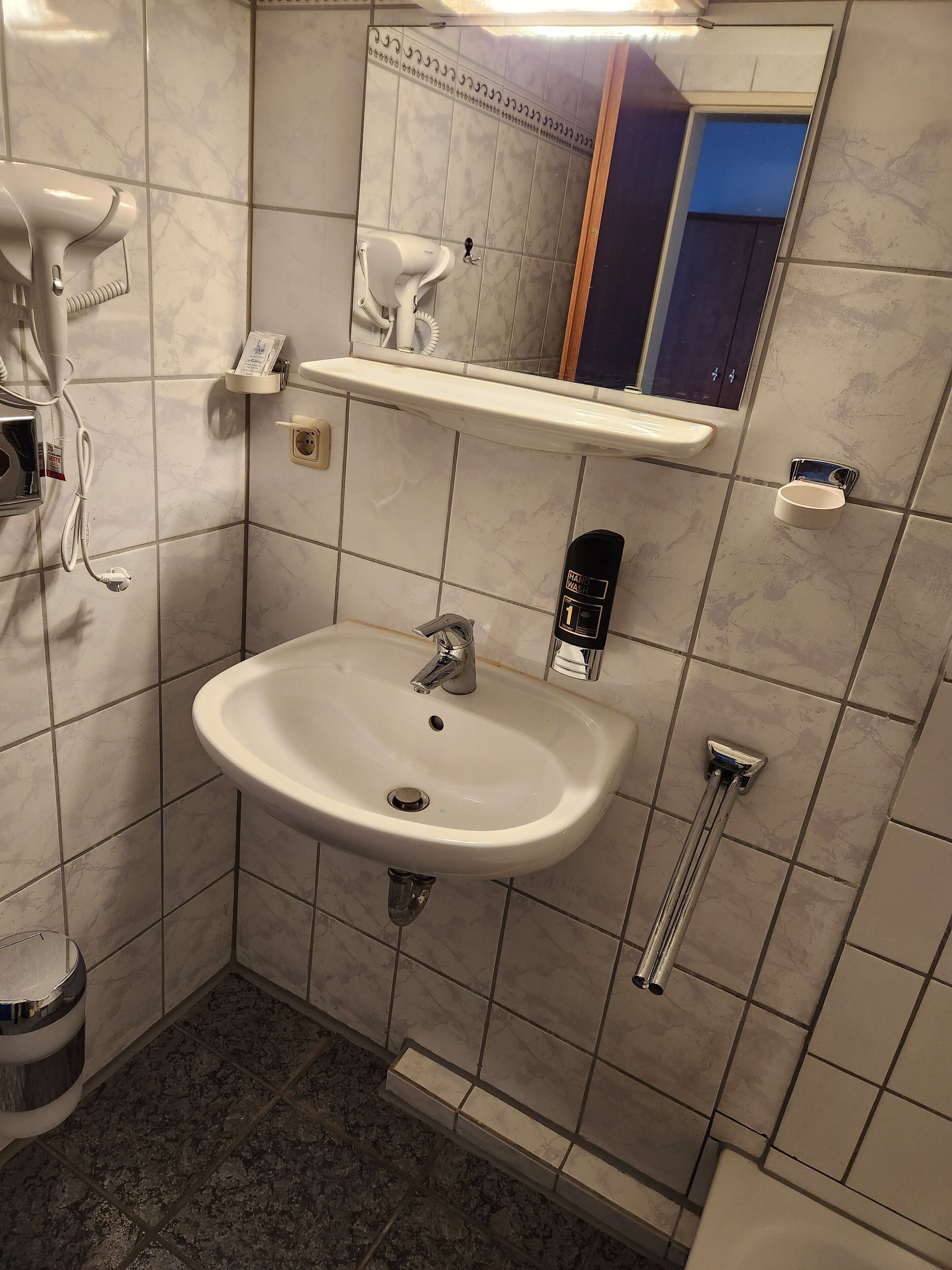 Ein Badezimmer mit einem weißen Waschbecken, einem Spiegel, einem Handtuchhalter und einem Händetrockner.