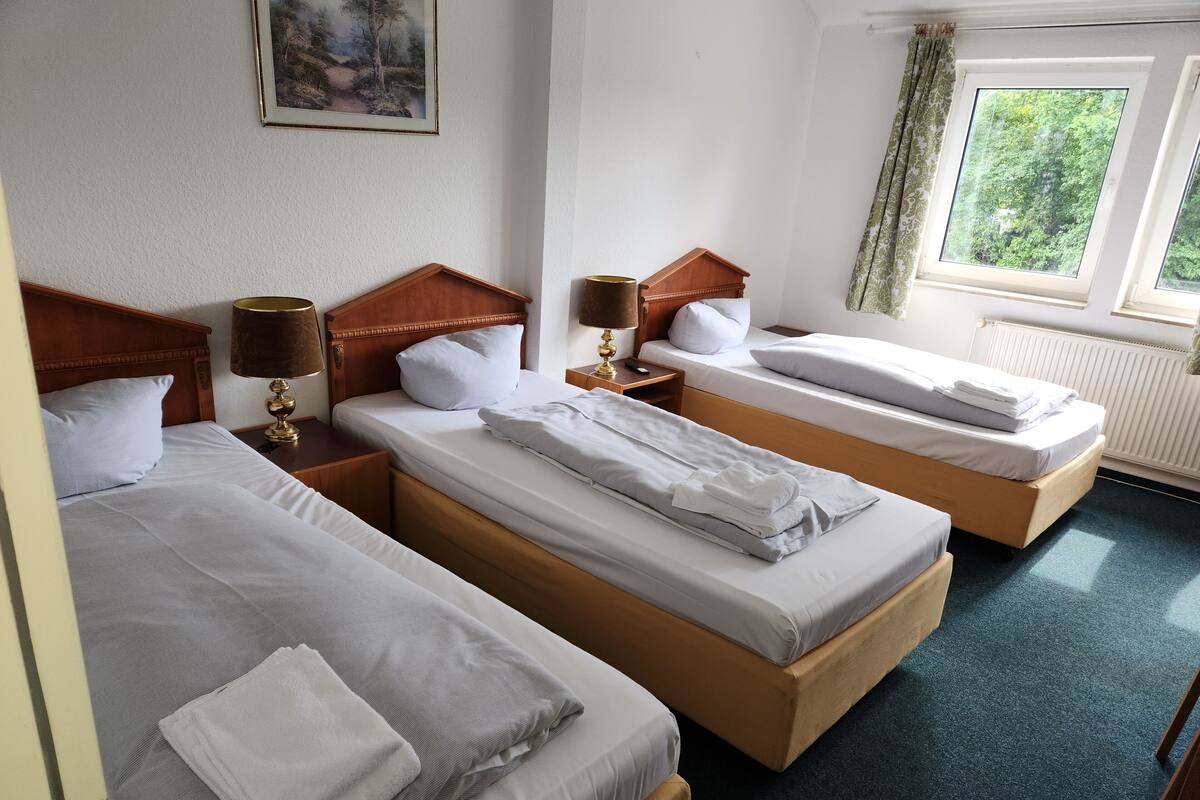 A hotel room with three single beds, each with a wooden headboard, white linens, and a small bedside table with a lamp. A painting is on the wall, and there is a window with curtains.
