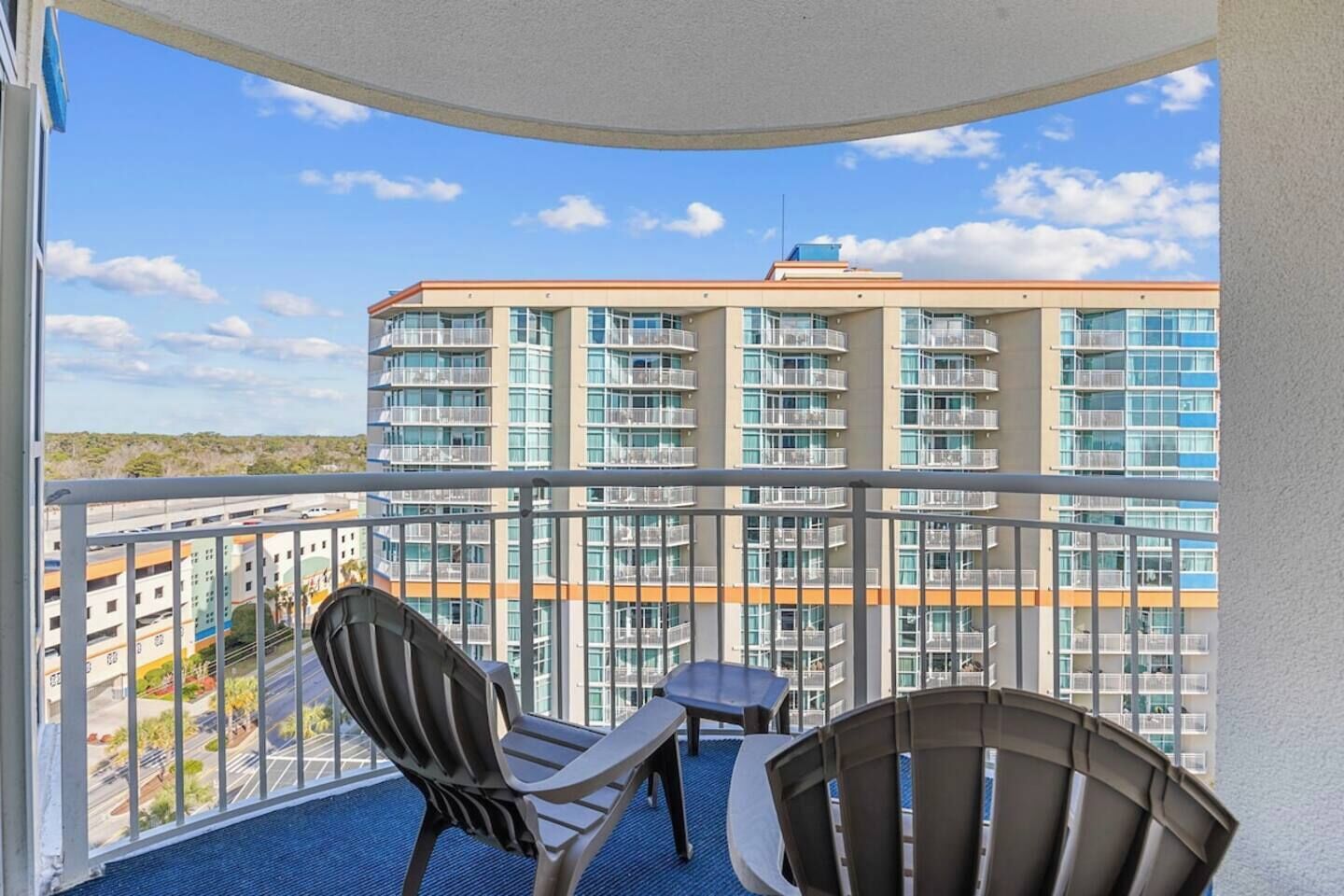 A balcony with two chairs and a small table, overlooking a high-rise building with multiple balconies.