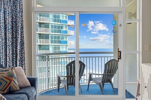A balcony with two Adirondack chairs, a blue patterned curtain, and a view of a building with balconies.