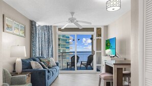A room with a blue sofa, a lamp, a TV, and a balcony with a view of the ocean.
