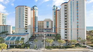 A multi-story hotel with a central entrance, parking area, and a view of the ocean.