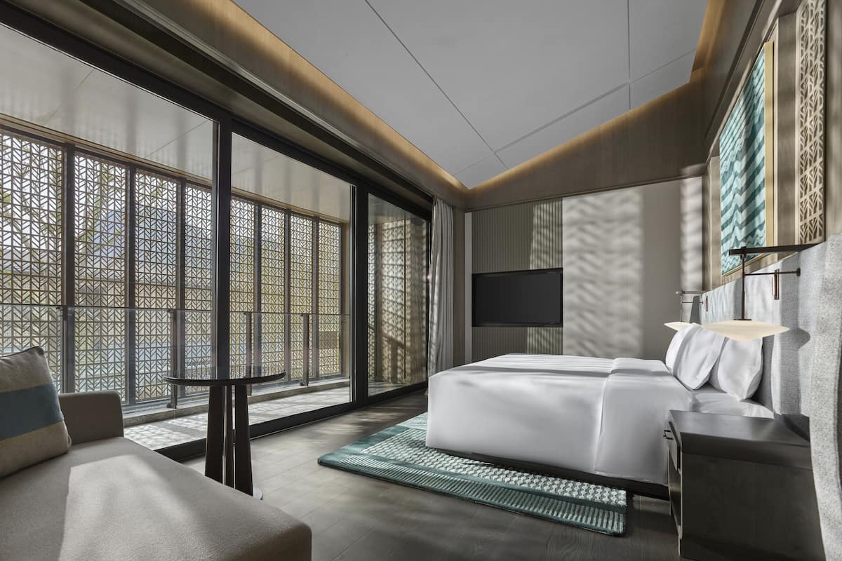 A modern hotel room with a large bed, a sofa, a TV mounted on the wall, and large windows with a view of the outside.