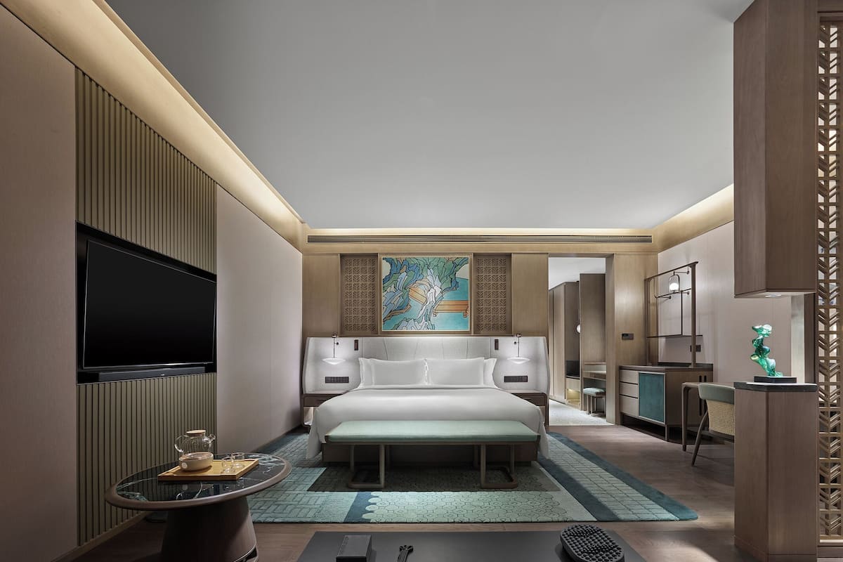 A modern hotel room with a large bed, a flat-screen TV, a desk, and a seating area with a small table and chair.