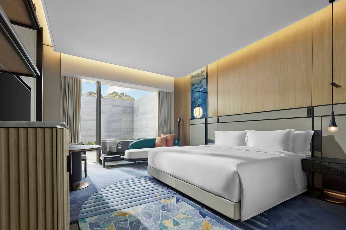 Una habitación de hotel moderna con una cama grande, una zona de estar con una mesa y sillas, un ventanal con vistas al exterior y un televisor montado en la pared.