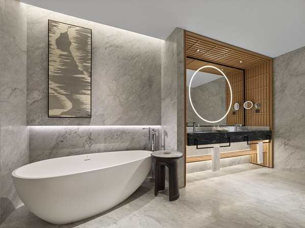 A modern bathroom with a freestanding bathtub, a wooden vanity with a large mirror, and a marble wall. - Angsana Suzhou Shishan (Suzhou)