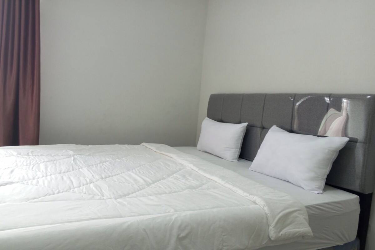 A neatly made bed with white bedding and two pillows, a grey upholstered headboard, and a plain wall with a brown curtain to the left.