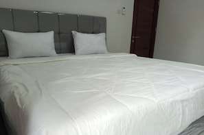Cama bem arrumada com cabeceira cinza e roupa de cama branca.