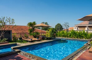 A swimming pool with lounge chairs and umbrellas, surrounded by greenery and a tiled roof building in the background. - SIEM REAP RIVER VIEW RESIDENCE (Siem Reap)