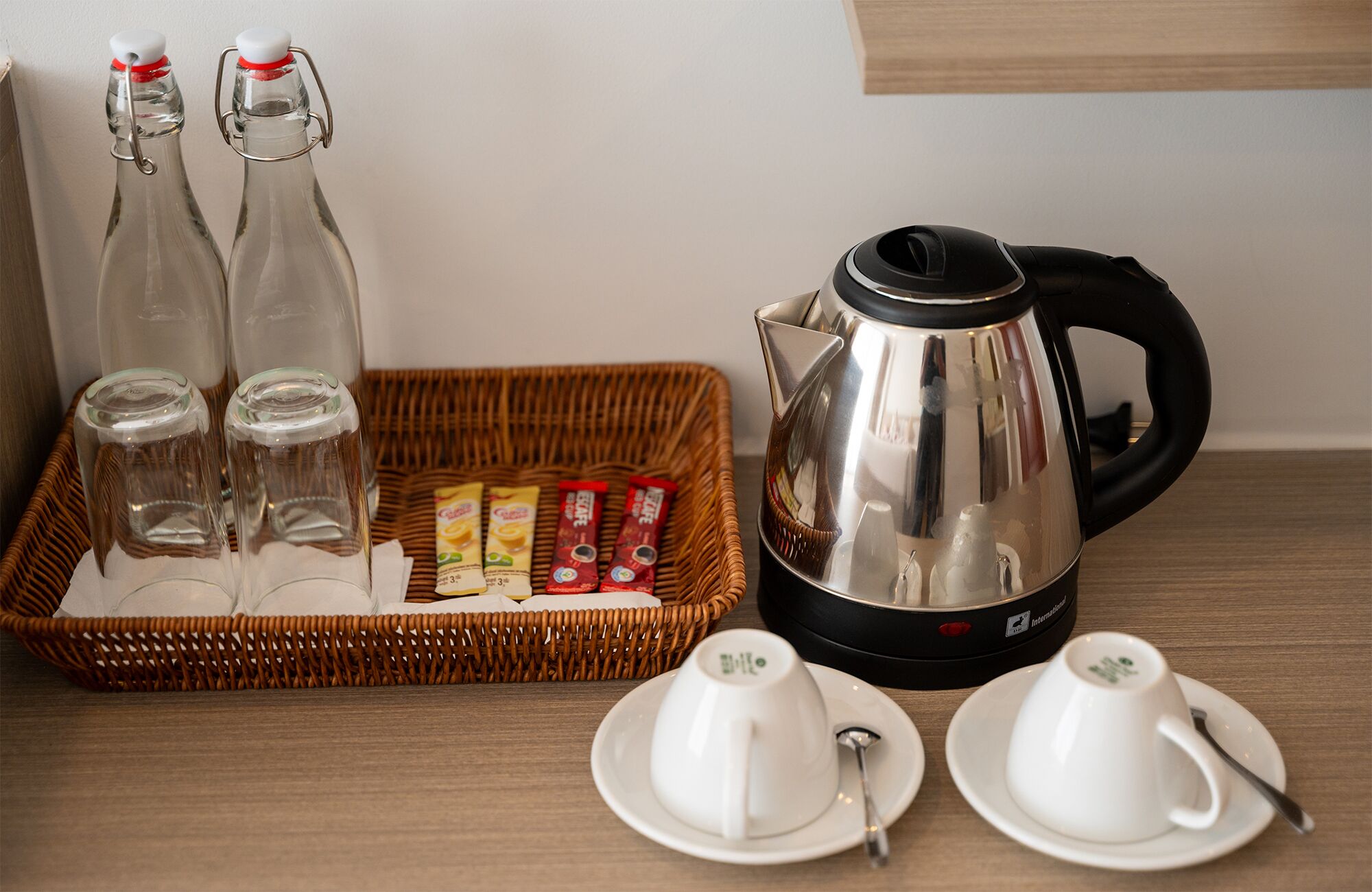 A breakfast setup with a kettle, two cups, a basket with packets, and two bottles.