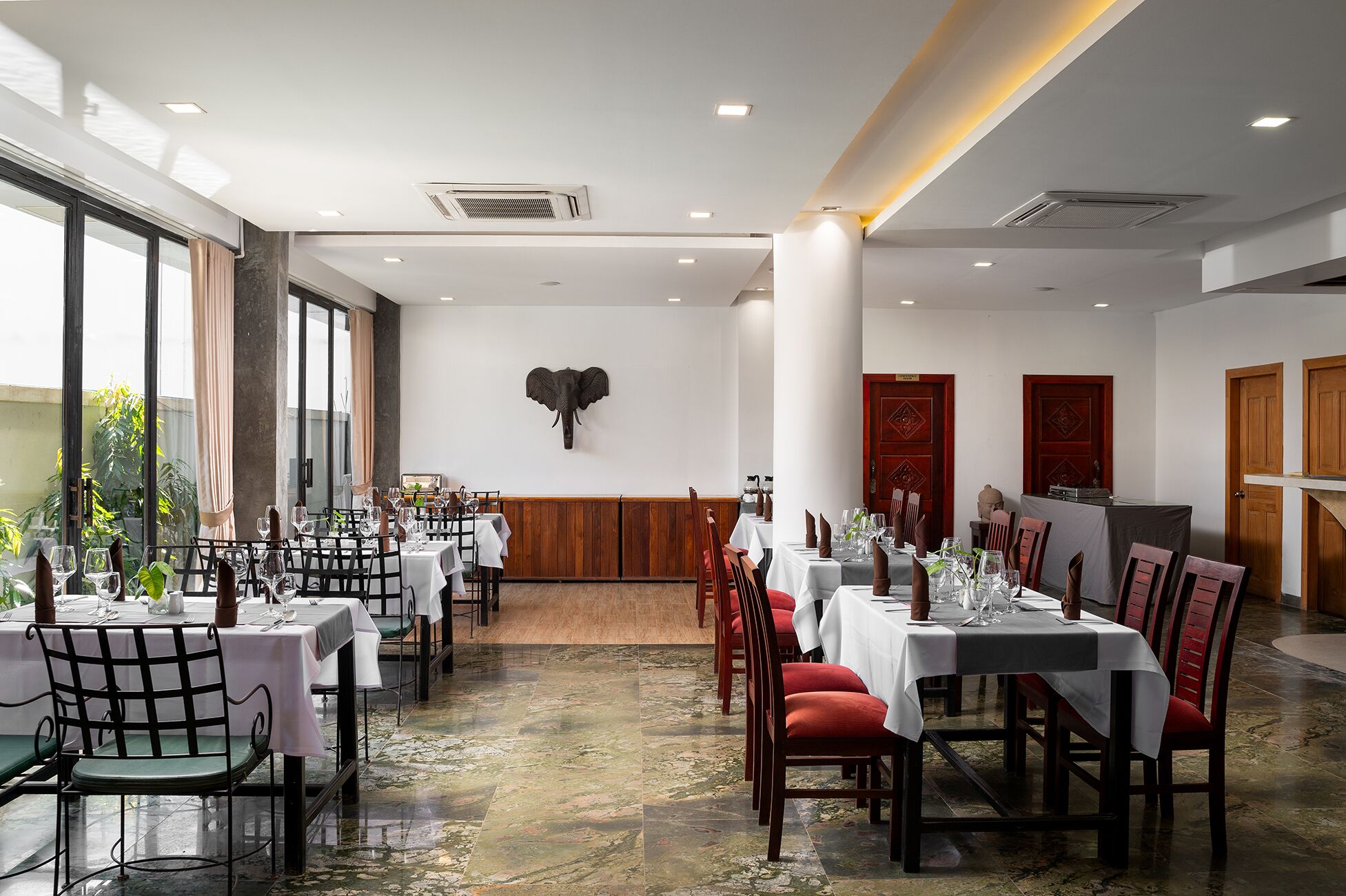 A restaurant with multiple tables and chairs, a large elephant sculpture on the wall, and wooden doors.