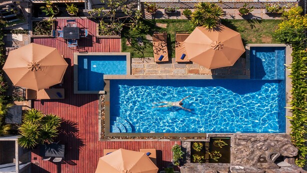 A rectangular swimming pool with a swimmer, surrounded by lounge chairs and umbrellas, set against a red deck with a stone wall and greenery. - SIEM REAP RIVER VIEW RESIDENCE (Siem Reap)