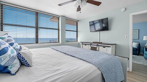 Une chambre à coucher avec un lit, un ventilateur de plafond, une télévision et une vue sur l’océan grâce à de grandes fenêtres.