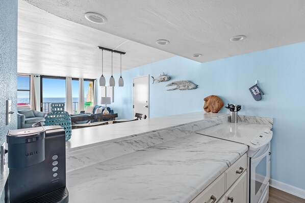 A modern kitchen with a marble countertop, a coffee maker, and a view of the ocean through large windows.