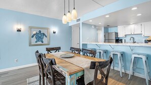 A dining area with a wooden table and chairs, a light blue wall, and a kitchen with white cabinets and a stainless steel refrigerator. - Beachfront Gulf House, 602, Sea South Properties (Gulf Shores)