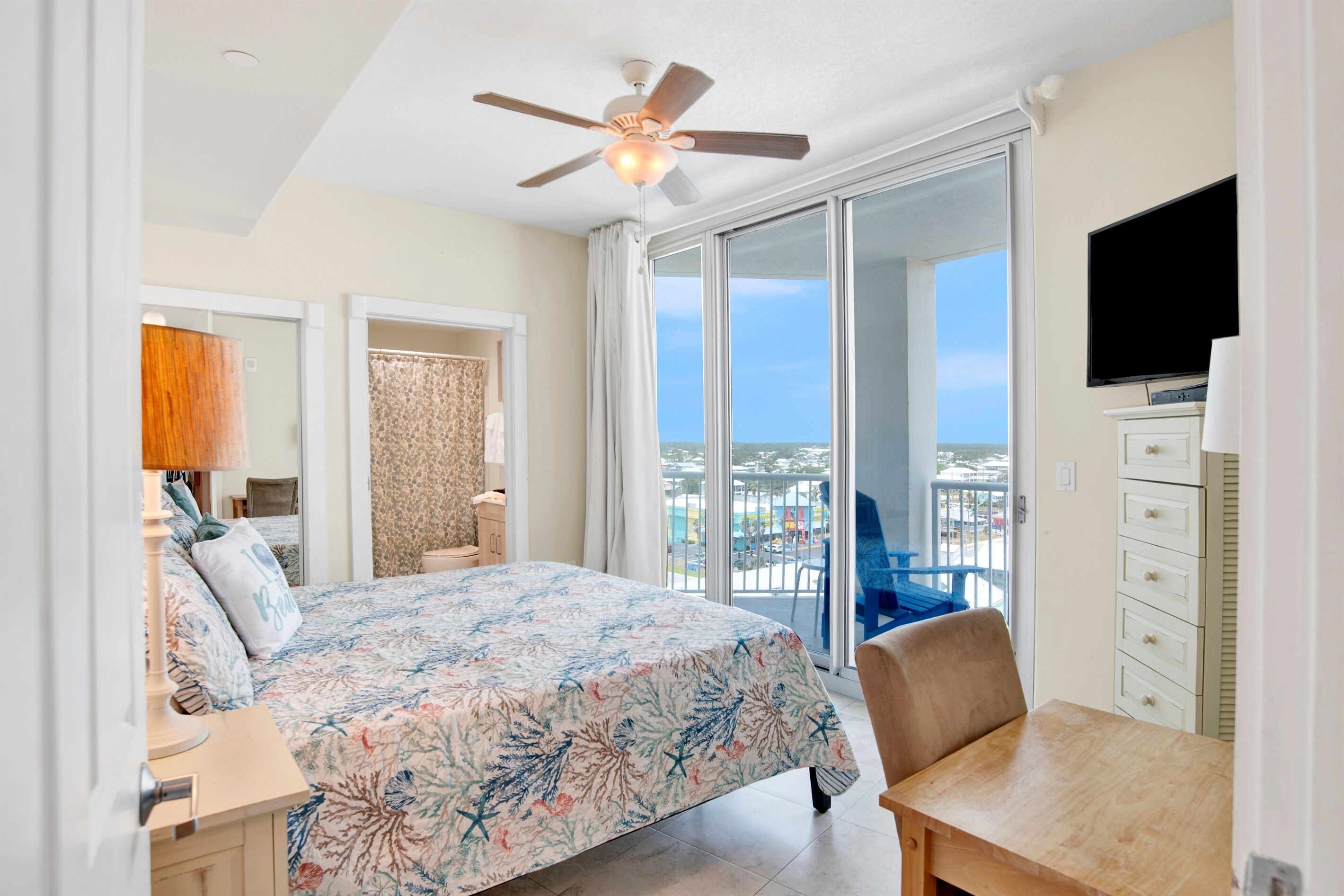 A bedroom with a bed, a desk, a chair, a TV, and a balcony with a view.