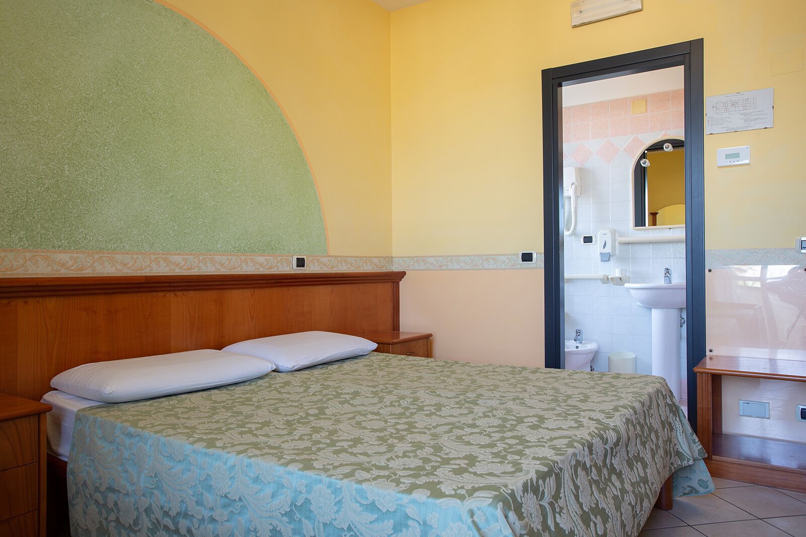 Een slaapkamer met een bed, een houten hoofdbord, een nachtkastje en een zichtbare badkamer door een open deur.