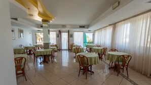 Un comedor con mesas y sillas redondas, ventanas amplias con cortinas y piso de baldosas.