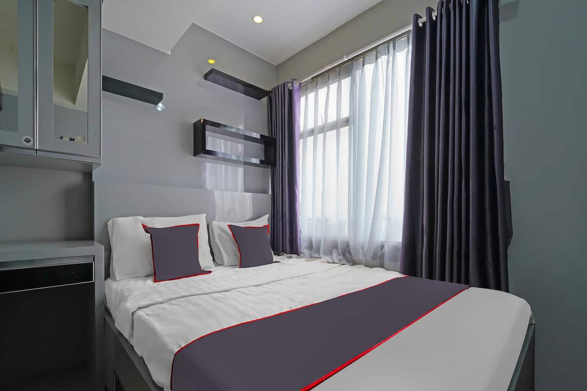 A modern hotel room with a large bed, grey and white bedding, a small bedside table, and a window with sheer curtains.