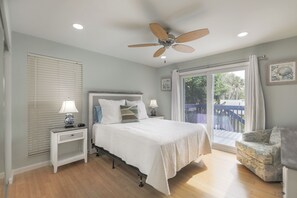 A bedroom with a bed, nightstands, a ceiling fan, a window with blinds, a balcony with a view, and a chair.