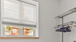 Een kleine kamer met een raam, een houten plank, twee krukken en een metalen rek met kleding.