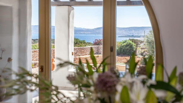 Blick aus einem Zimmer mit einem großen Rundbogenfenster auf eine Küstenlandschaft mit Bergen und blauem Himmel.