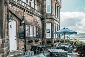 The exterior of Victoria Hotel with stone facade, outdoor seating, and a view of the sea.