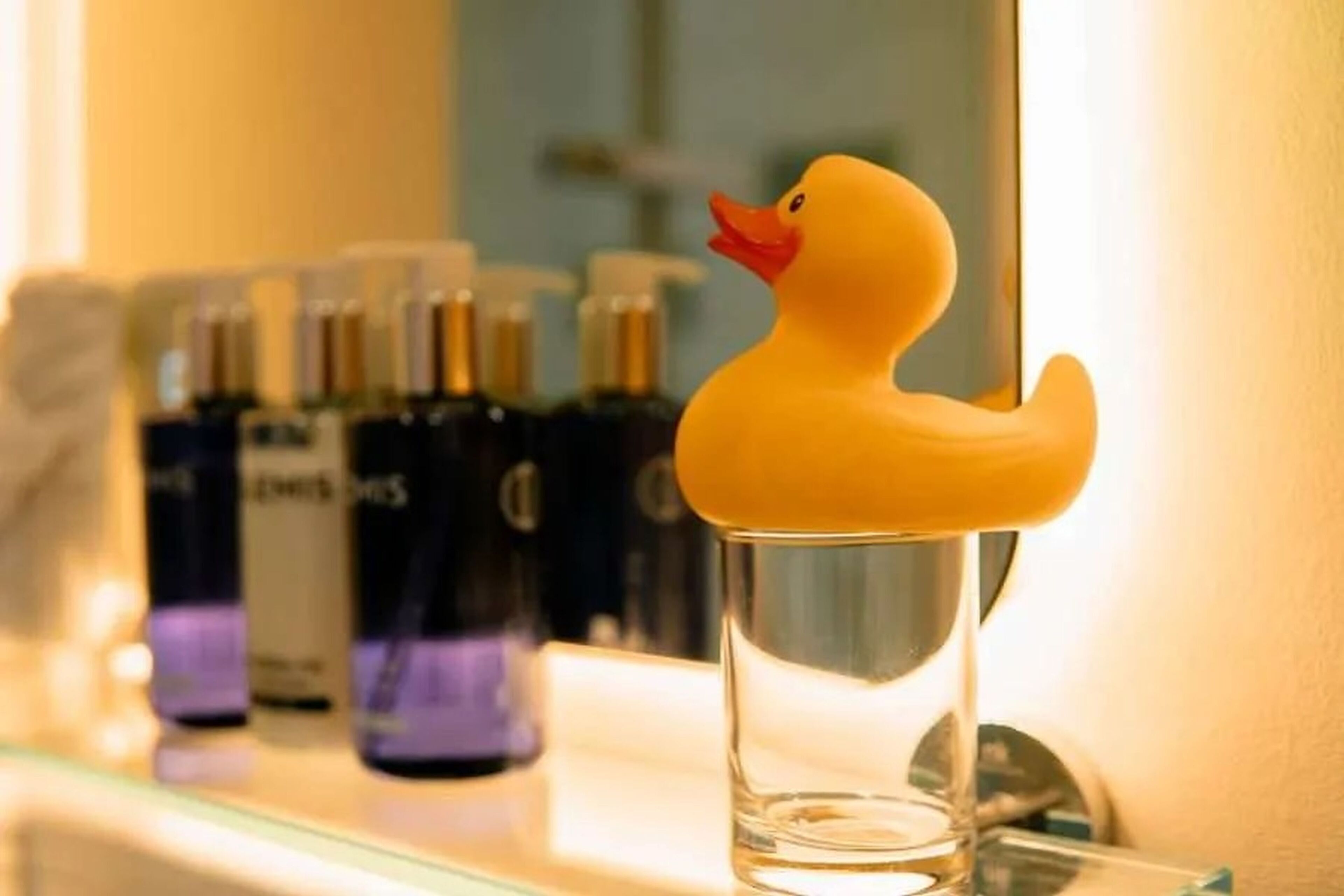 A clear glass of water with a rubber duck on the rim, placed on a shelf with various bottles in the background.