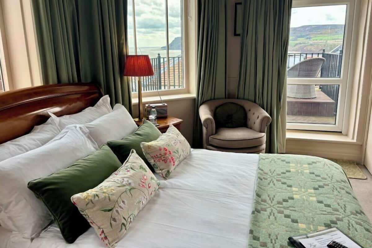 A hotel room with a bed, a chair, a table with a lamp, and a view of the sea through large windows.