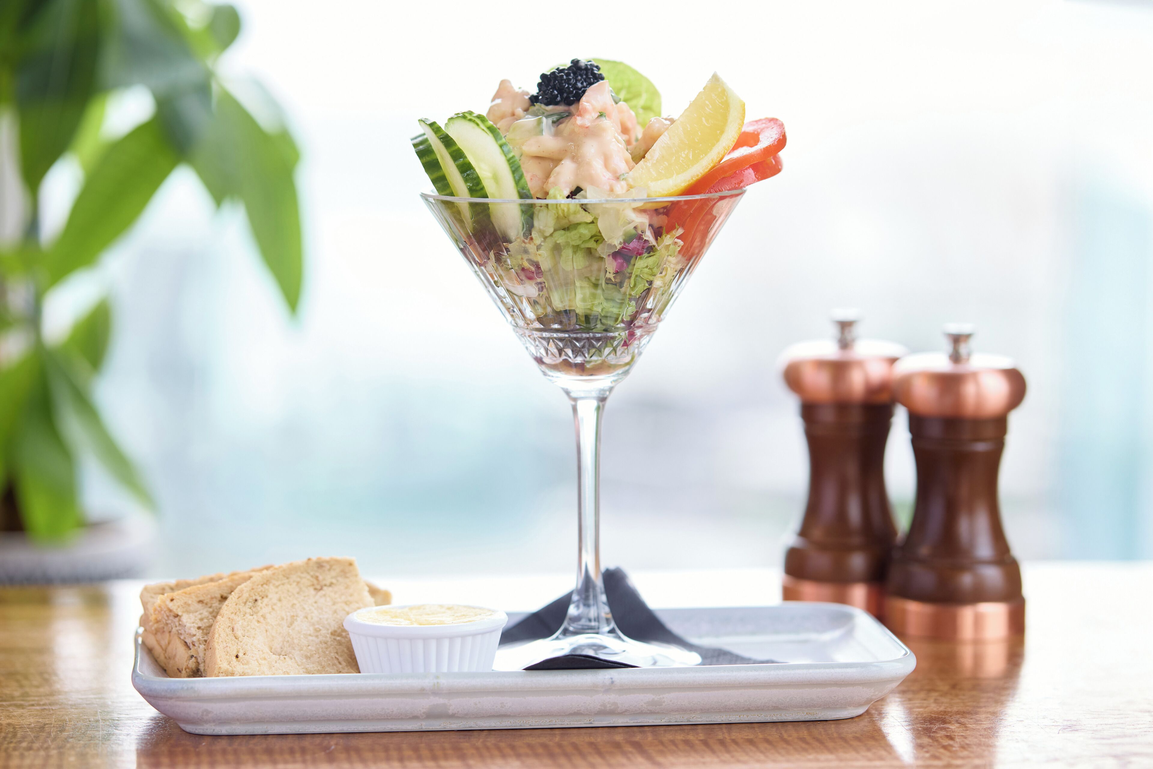 Salada numa taça de vidro com pão e um pequeno prato de molho sobre uma mesa de madeira.