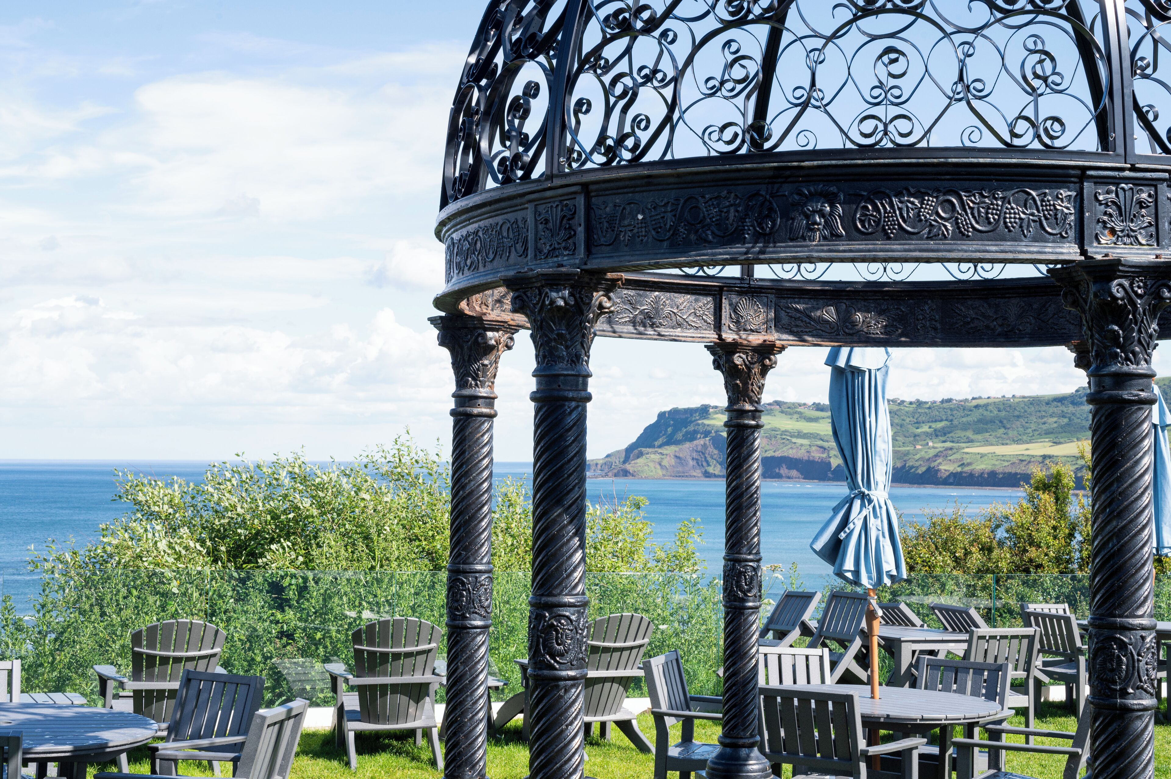 A gazebo with intricate metalwork, surrounded by outdoor seating and overlooking a scenic view of the sea and hills.