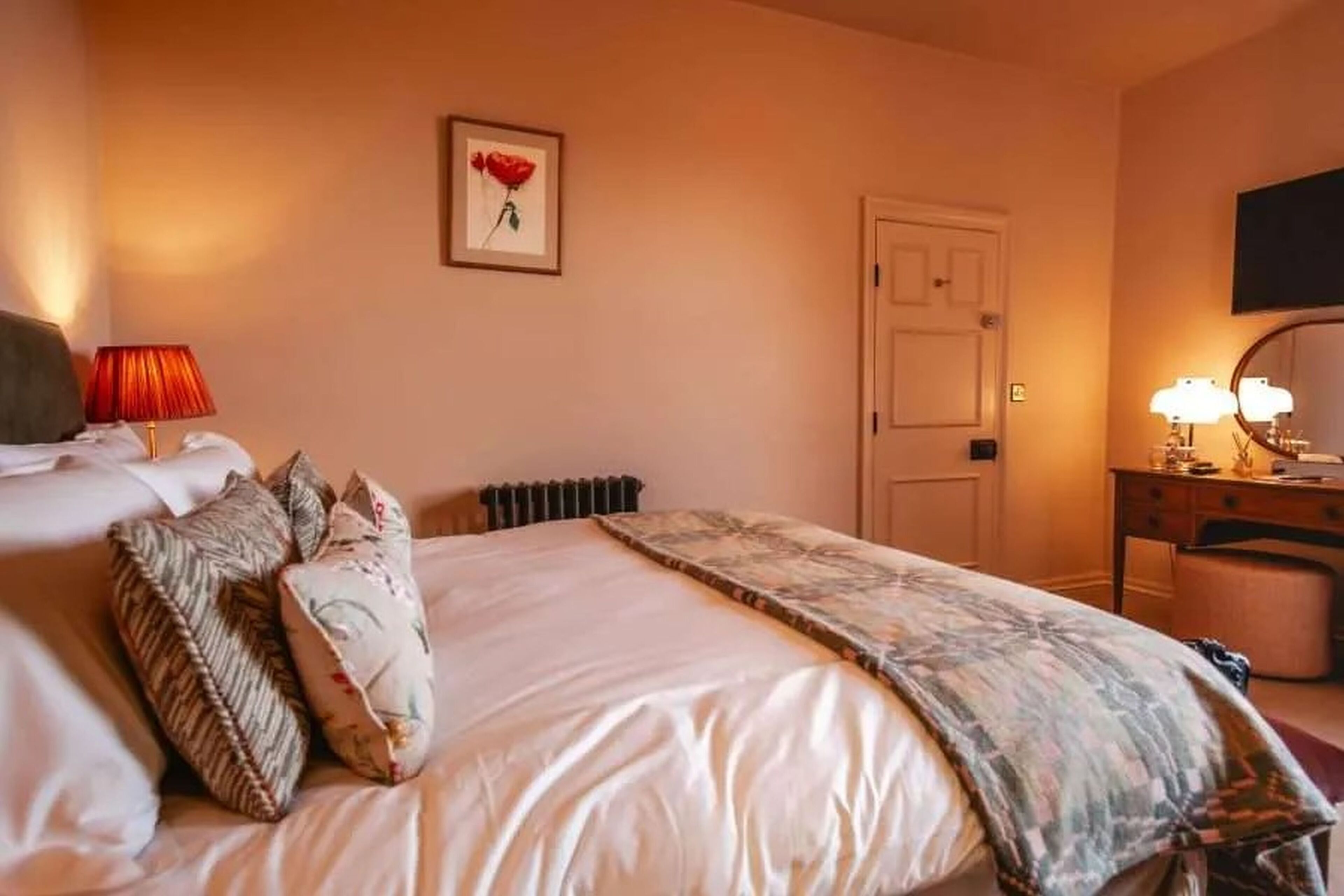 A hotel room with a bed, a desk with a lamp, a chair, a radiator, and a door.