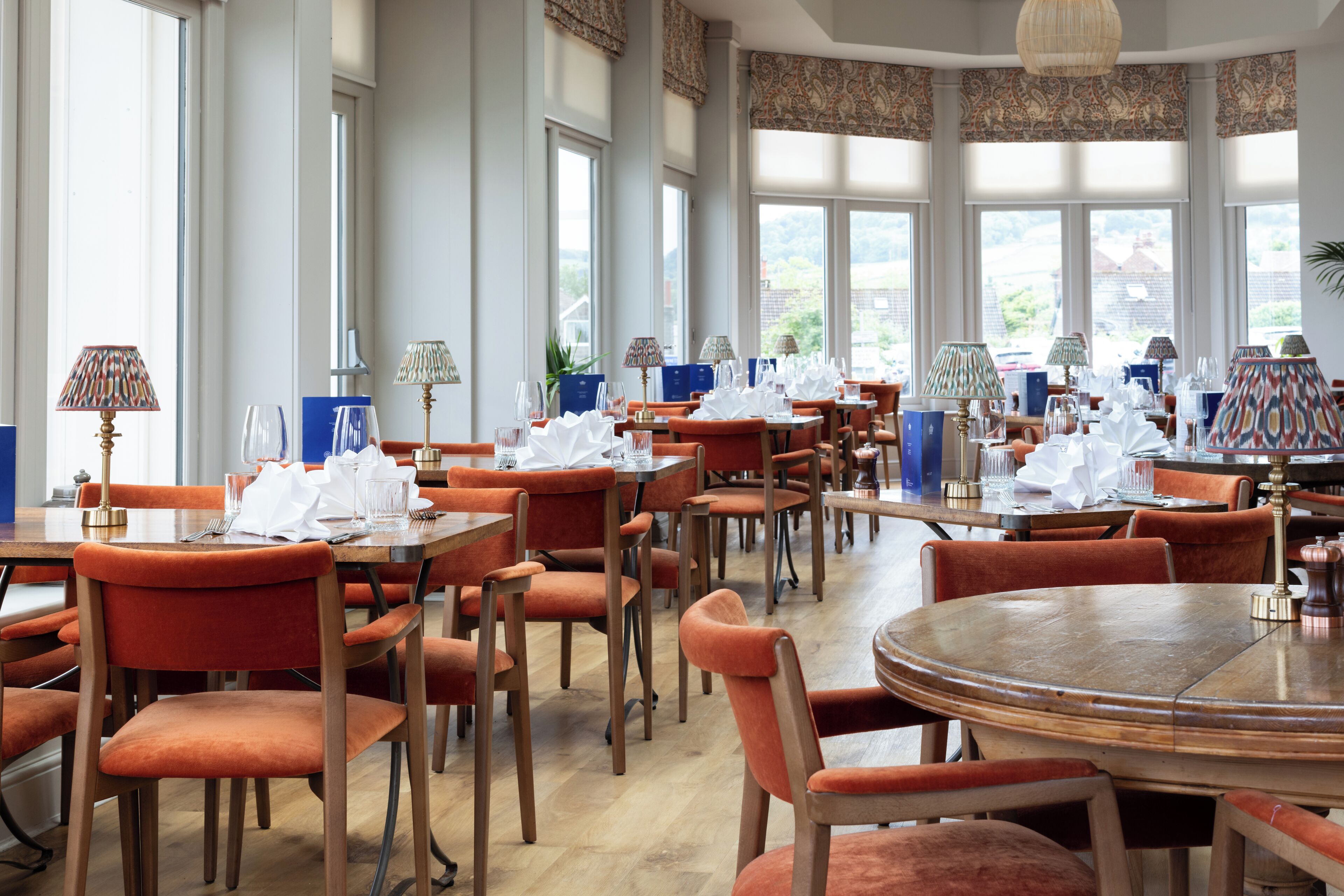 A restaurant with multiple tables and chairs, large windows, and patterned curtains.