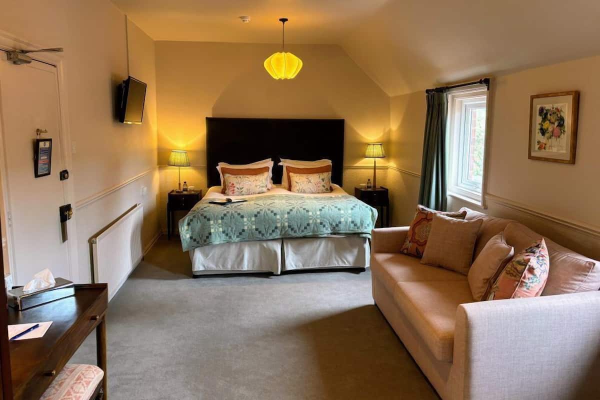 A bedroom with a bed, two bedside tables, a nightstand, a sofa, a desk, a chair, a radiator, a ceiling light, and a window with curtains.