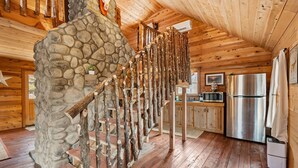 Interior rústico de uma cabana com escadaria de pedra, paredes de madeira, área de cozinha e uma geladeira grande.