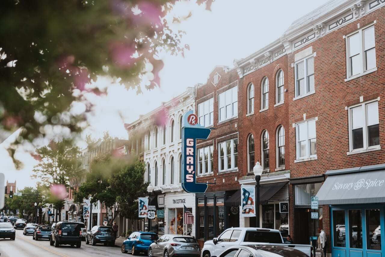 A street view with a row of brick buildings, a pharmacy sign, and a Tin Cottage store.