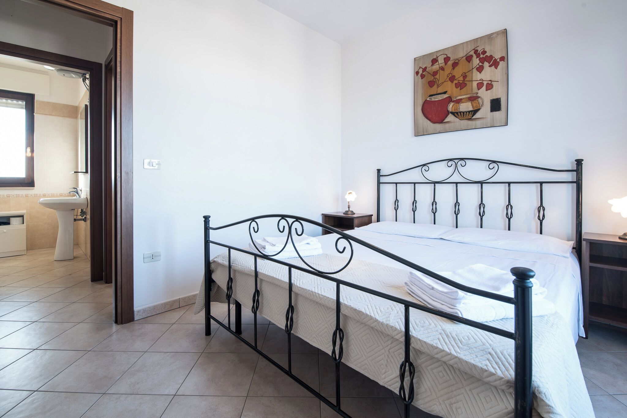 Une chambre à coucher avec un lit en métal, du linge de lit blanc, une table de chevet avec une lampe et un tableau accroché au mur.