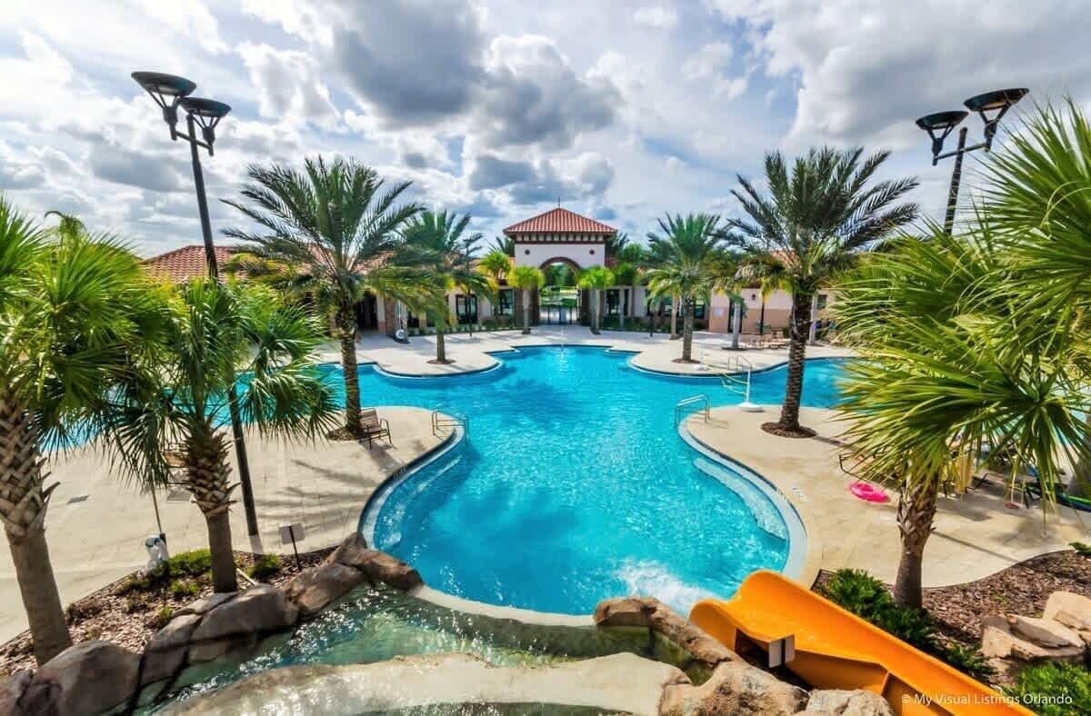 A swimming pool with a slide, surrounded by palm trees and a building with a red roof in the background.