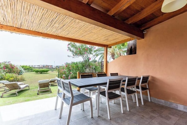 An outdoor patio with a table and chairs, a thatched roof, and a view of a garden.