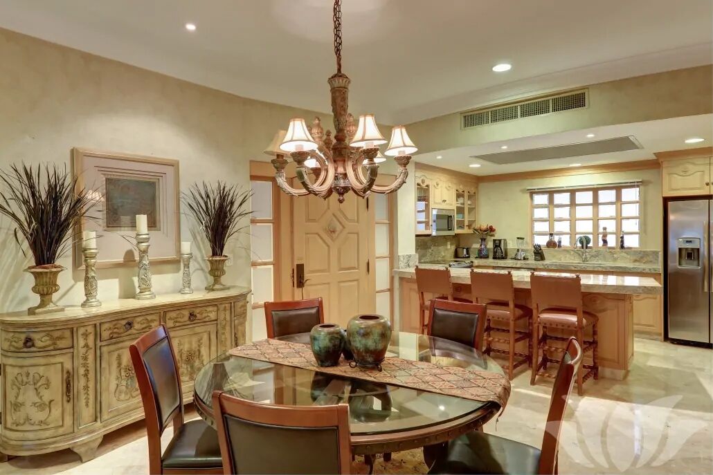 A dining area with a glass table, chairs, a chandelier, and a kitchen in the background.